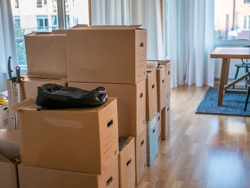 Stacks of cardboard moving boxes with an empty backpack on top at a home with wooden floors.
