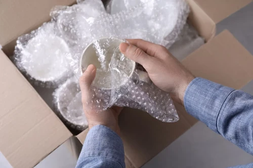 A person packing glassware into a cardboard box lined with Bubble Wrap.
