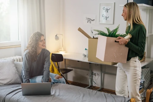 Friends moving into an apartment. one holding a box and the other typing on the bed.