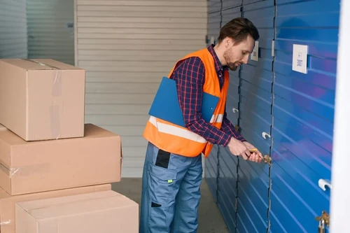 A person locking a blue storage unit door with moving boxes stacked nearby.