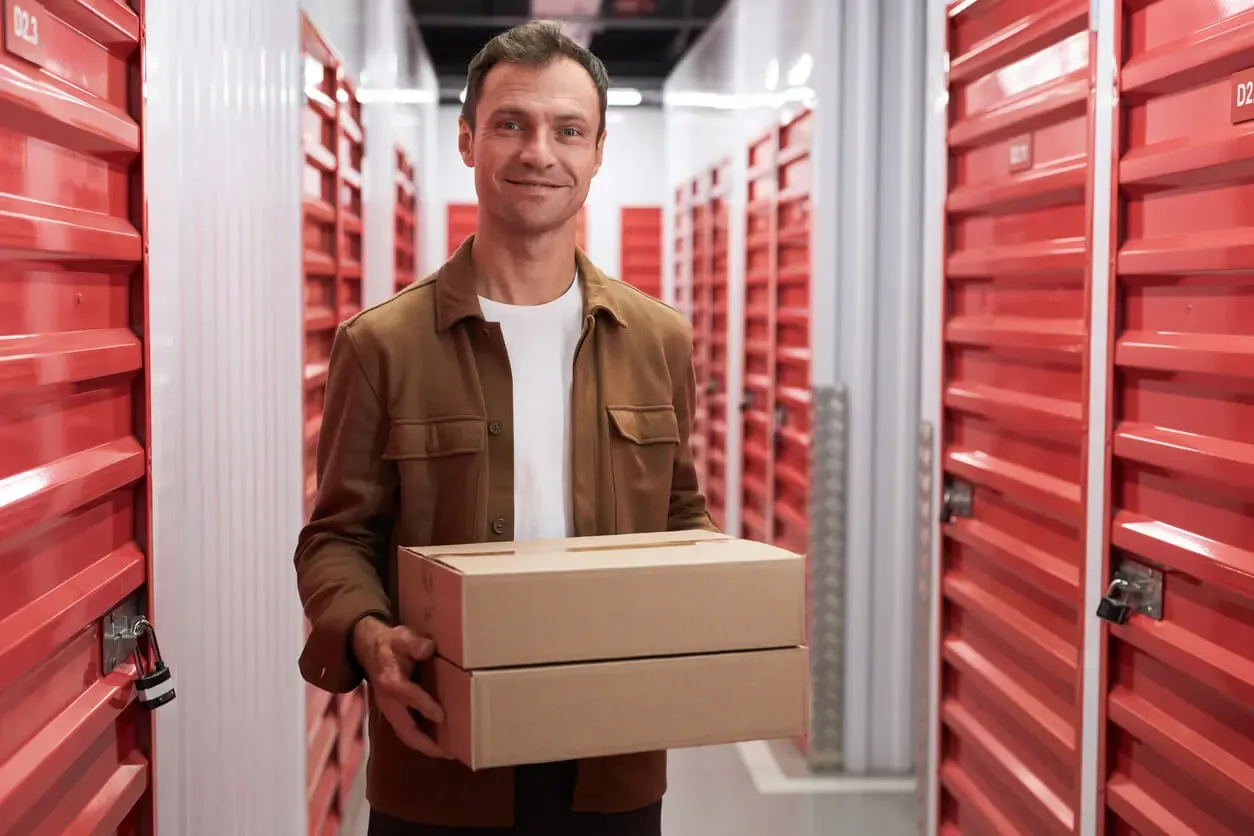 Man carrying boxes inside indoor self storage hallway with red storage unit doors.