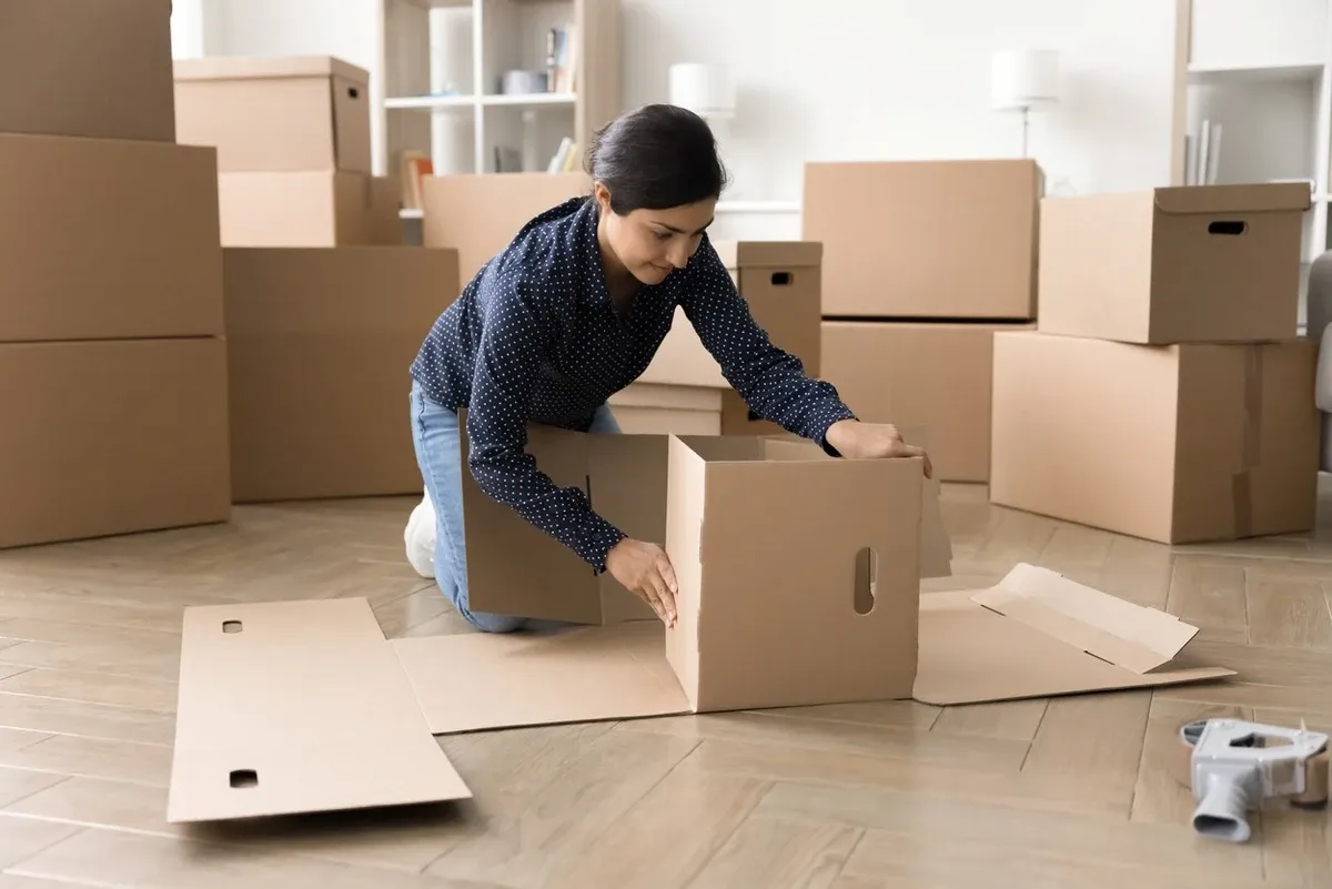 A young woman folds cardboard boxes into shape as she’s surrounded by other boxes in her living room.