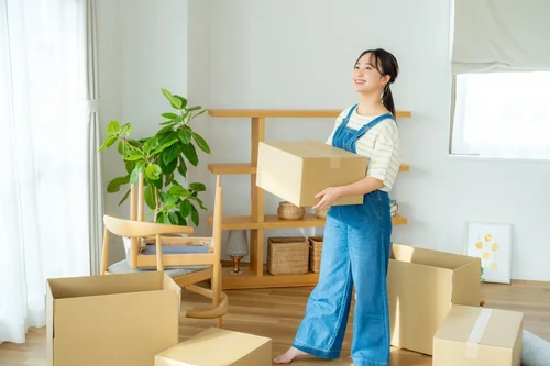 A smiling young woman carrying a cardboard box looks around her apartment, which is filled with moving boxes.